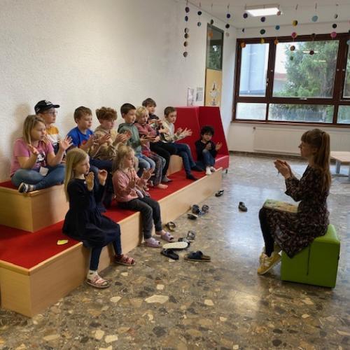 eine Gruppe Kinder sitzt auf einer roten Sitzstufe. Die Lehrerin sitzt gegenüber auf einem gründen Würfel  - alle klatschen