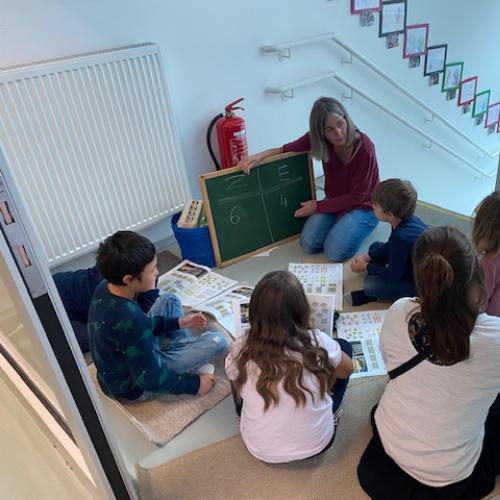 Kinder sitzen im Stiegenhaus beim Rechnen mit Lehrerin und "Hand"-Tafel