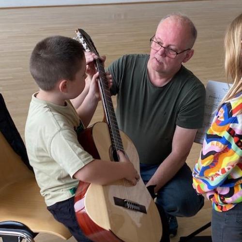 Kind sitzt mit viel zu großer Gitarre am Stuhl - der Lehrer hilft