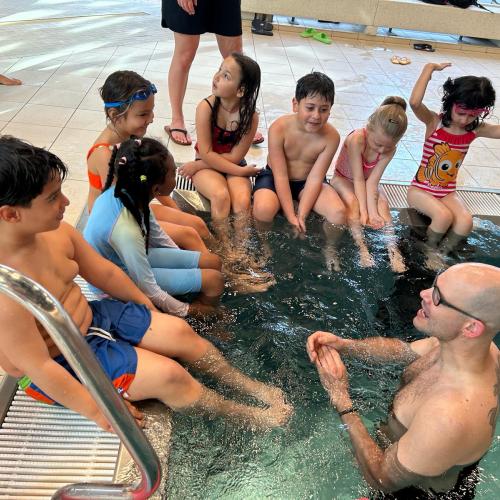 Kinder sitzen am Beckenrand und sprechen mit dem Schwimmlehrer