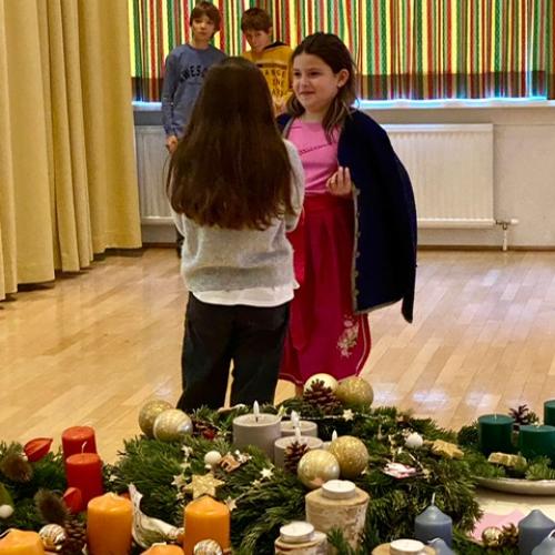 Man sieht im Vordergrund Adventkränze und dahinter Kinder bei der Aufführung