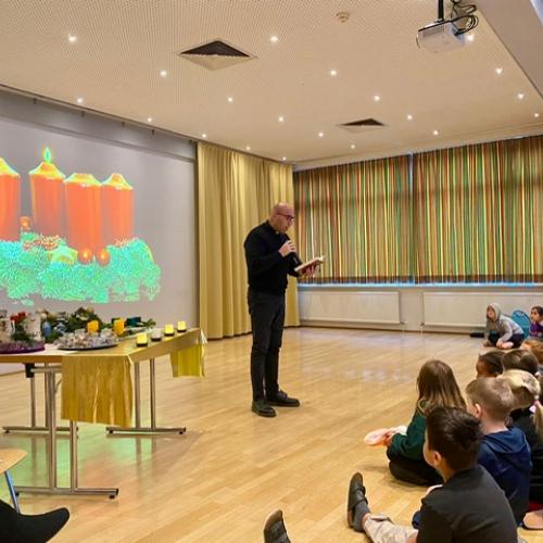Pfarrer Gimmy steht vor den Kindern, dahinter ein großes Bild eines Adventkranzes