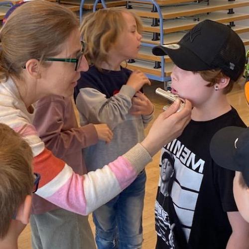 Kind mit Mütze bläst gerade in den Flötenkopf, den die Lehrerin hinhält