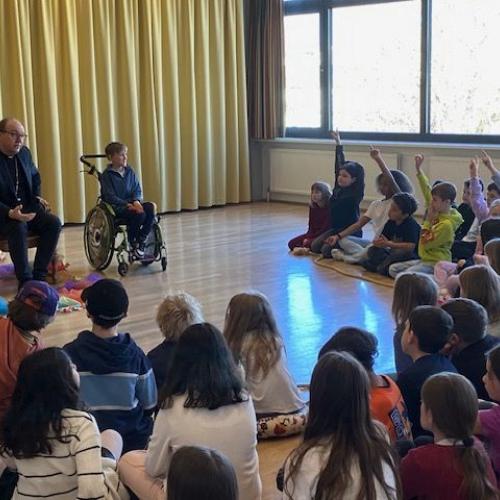In Vordergrund sitzen die Kinder im Halbkreis - im linken Bildrand sitzt der Bischof, daneben ein Kind im Rollstuhl
