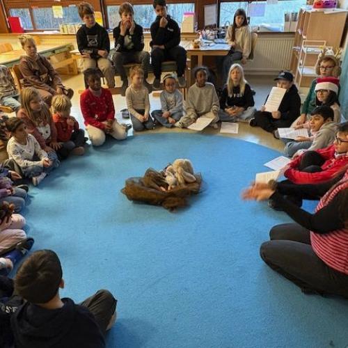 Die Kinder sitzen im Kreis am Boden. Die Lehrerin erklärt gerade. In der Mitte liegt ein Tuch mit versch. Materialien