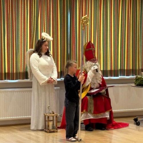 Engel steht neben dem sitzenden Nikolaus. Davor steht ein Mädchen, das den Stab hält