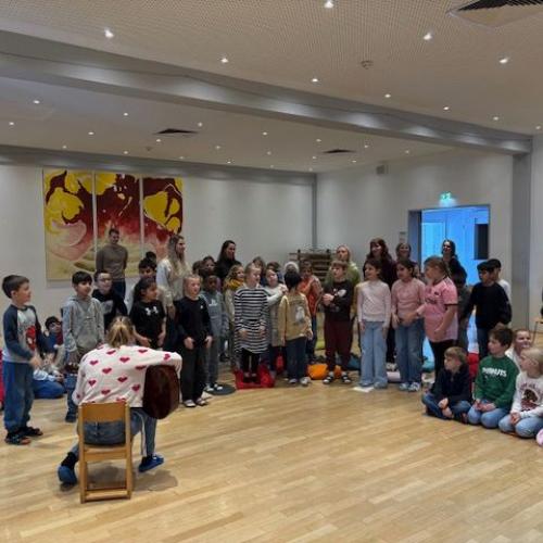 Lehrerin sitzt mit dem Rücken zum Fotografen - mit Gitarre in der Hand. Im Hintergrund eine Klasse, die ein Nikolauslied singt