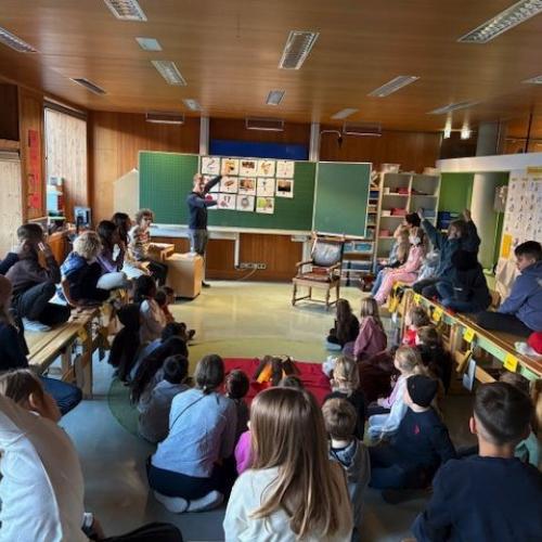 Lehrer erklärt Begriffe anhand von Bildern an der Tafel -rundum sitzen Kinder auf dem Boden und den Tischen
