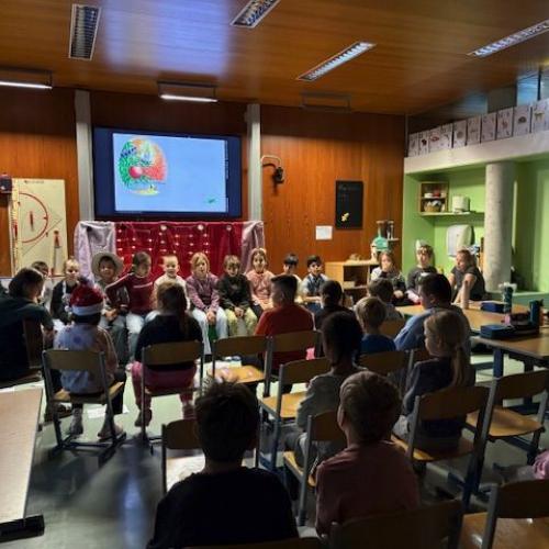 Lehrerin spielt Gitarre, große Kinder sitzen im Kreis und singen, während die Kleinen zusehen
