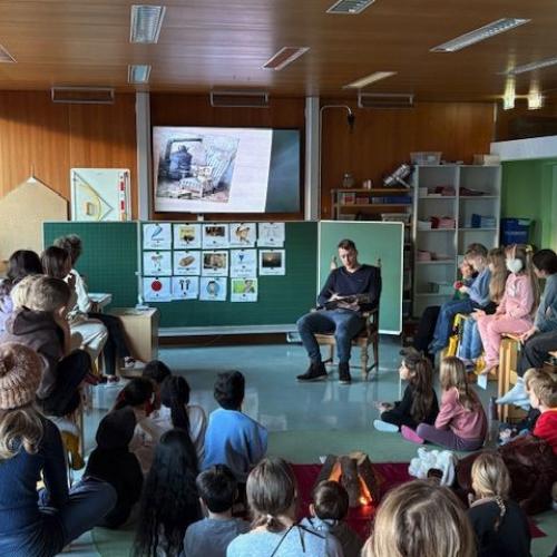 Lehrer sitzt vorne und liest ein Bilderbuch; Kinder sitzen im Kreis rund um ihn; im Hintergrund sieht man die Bilderbuchseite am Bildschirm