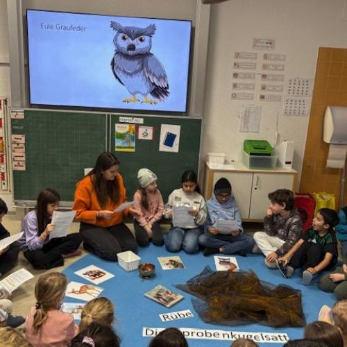 Lehrerin sitzt im Sitzkreis mit den Kindern, im Hintergrund sieht man am Bildschirm eine Eule