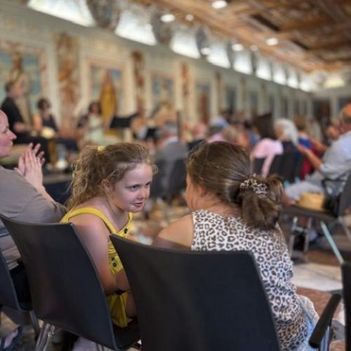 2 Kinder unterhalten sich; im Hintergrund sieht man viele sitzende Menschen im spanischen Saal