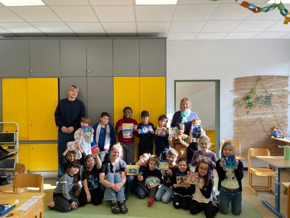 Eine Schulklasse mit rund zwanzig Kindern steht und sitzt gemeinsam in einem hellen Klassenzimmer. Die Kinder halten bemalte Holzplatten mit bunten Naturfarben in die Kamera. In der Mitte sitzen zwei erwachsene Frauen, links steht ein erwachsener Mann. Im Hintergrund sind gelbe und graue Schränke zu sehen. Die Kinder wirken stolz und fröhlich und präsentieren ihre kreativen Kunstwerke.