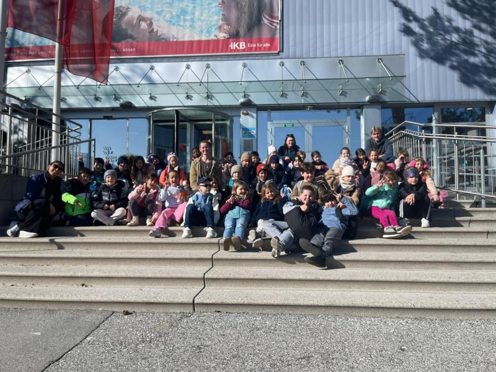 Kinder der Klassen 1a und 1b sitzen gemeinsam mit ihren Begleitpersonen auf den Stufen vor dem Eingang eines Hallenbads und posieren fröhlich für ein Gruppenfoto während ihrer Schwimmwoche. 