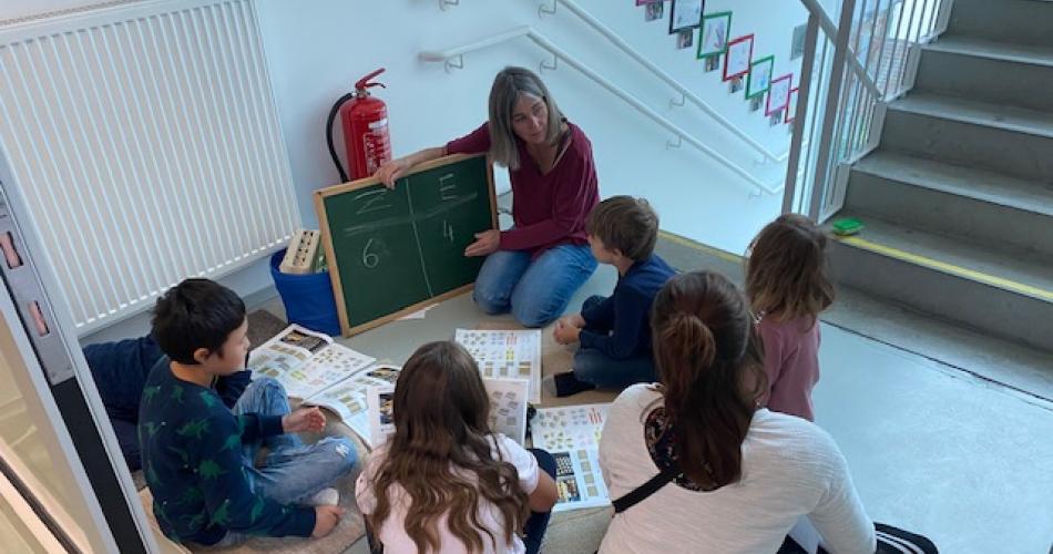 Kinder sitzen im Stiegenhaus mit Lehrerin + kleiner "Hand"-Tafel