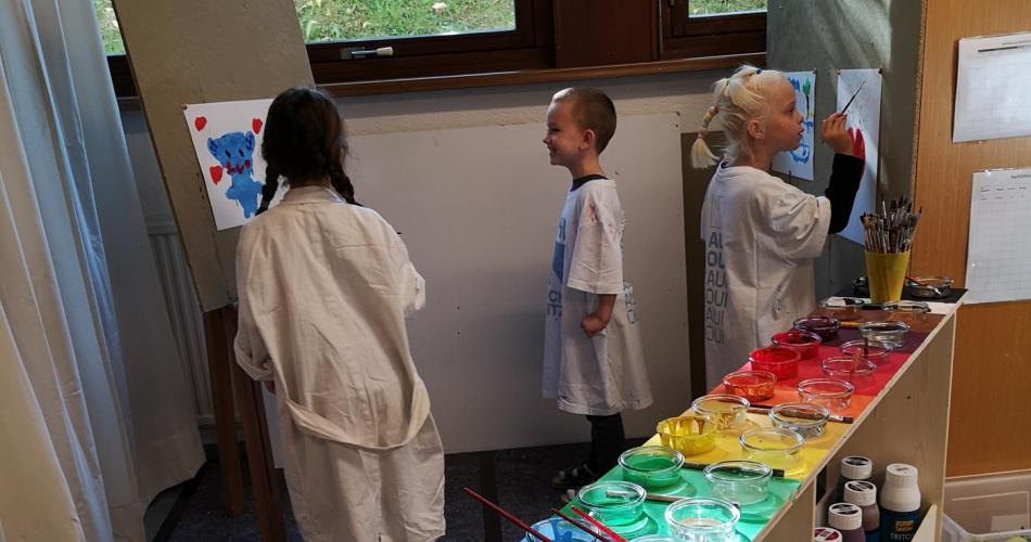 Kinder in weißen Malmanteln malen großflächig. Im Vordergrund steht ein Farbenregal