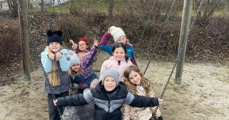 Acht Grundschulkinder sitzen und stehen gemeinsam auf einer großen Schaukel auf einem Spielplatz. Die Kinder tragen Winterjacken und Mützen und schauen lächelnd in die Kamera. Im Hintergrund sind Bäume, Sträucher und ein Gebäude zu sehen.