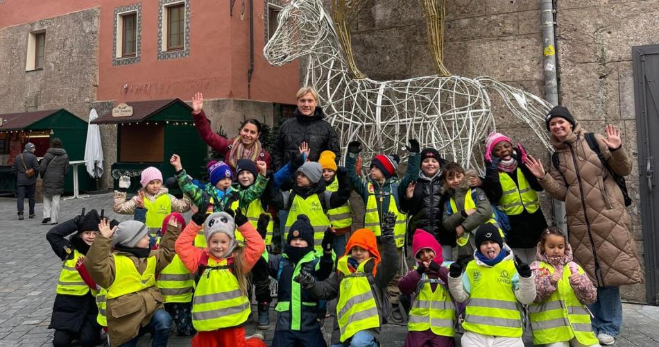 Eine Gruppe von Kindern in leuchtend gelben Sicherheitswesten posiert gemeinsam mit drei Erwachsenen für ein fröhliches Gruppenfoto auf einem Platz vor historischen Gebäuden. Hinter ihnen steht eine große, kunstvolle Drahtskulptur eines Reiters auf einem Pferd, weihnachtlich dekoriert.