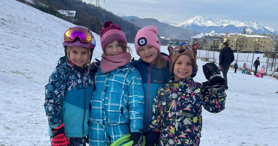 Vier Kinder stehen nebeneinander im Schnee und halten bunte Rutschblätter. Im Hintergrund sieht man Berge und weitere spielende Kinder.