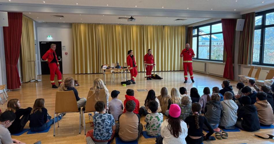 Rettungshundeführerinnen und -führer stellen ihre Ausrüstung vor und erklären den Kindern in der Aula die Arbeit der Rettungshundestaffel.