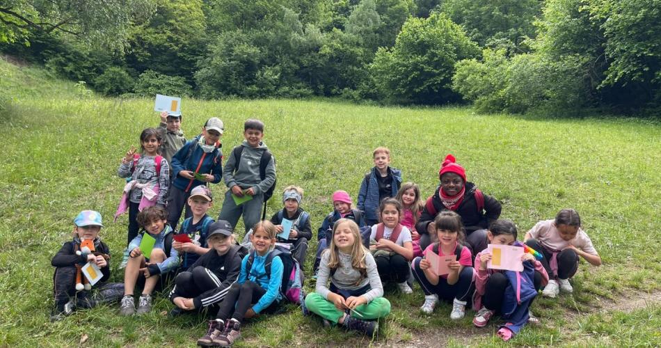 Kinder einer Schulklasse bei einer Exkursion auf einer Waldwiese mit Arbeitsmaterialien in den Händen.