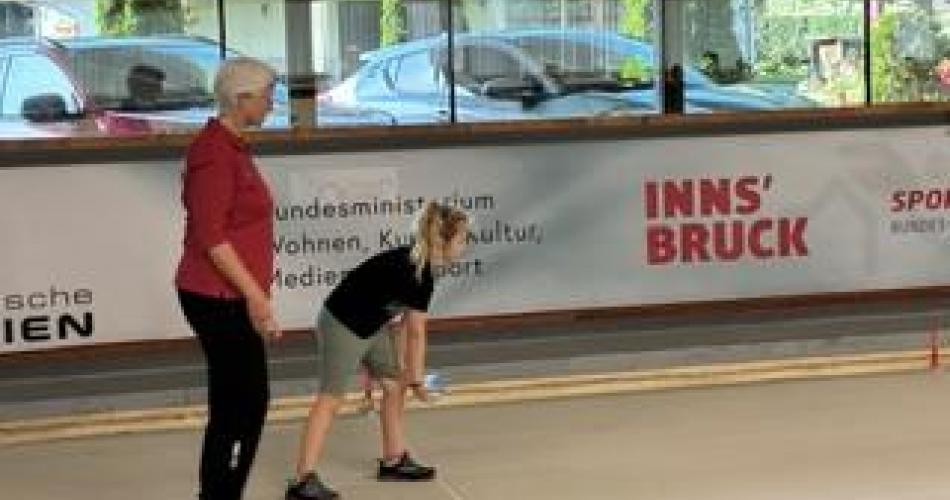 Kind beim Werfen der Boccia-Kugel; dahinter steht die Trainerin