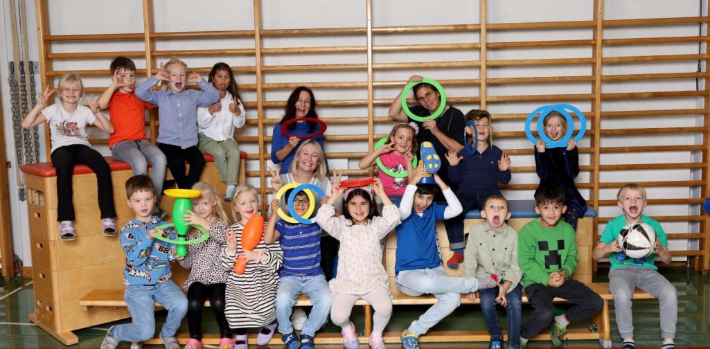 1a sitzt auf Langbänken und Kästen mit buntem Spielmaterial im Turnsaal.