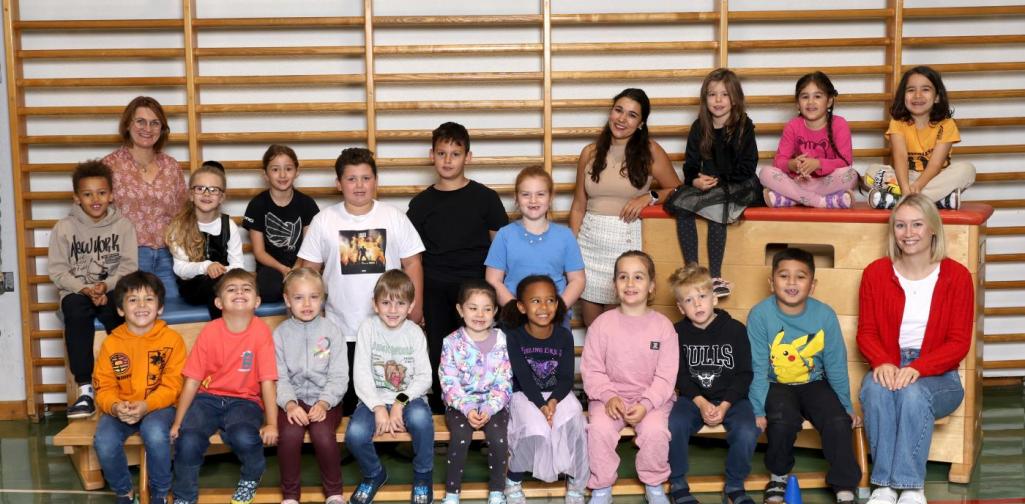 1b sitzt auf Langbänken und Kästen mit buntem Spielmaterial im Turnsaal.