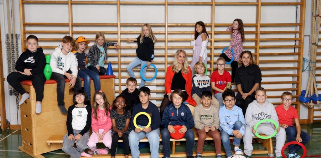 3a sitzt auf Langbänken und Kästen mit buntem Spielmaterial im Turnsaal.