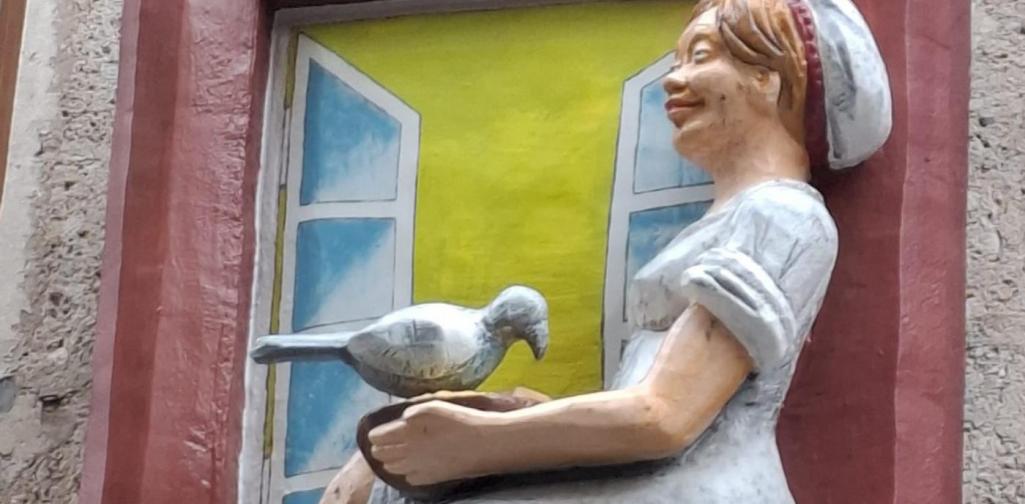 Alexandra Sigitz Eine Fassadenfigur stellt Aschenputtel dar: Sie sitzt barfuß in einem weißen Kleid auf einem roten Fensterbrett und füttert Tauben aus einer Schale. Zwei Tauben picken an ihrem Schoß, eine weitere sitzt oben auf dem Rahmen. Der Hintergrund zeigt gemalte, offene Fensterläden vor gelbem Licht.