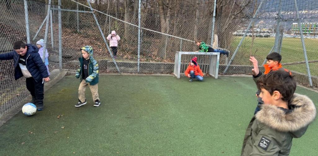 Mehrere Grundschulkinder spielen auf einem eingezäunten Sportplatz Fußball. Einige Kinder laufen dem Ball nach, andere stehen oder sitzen am Rand. Alle tragen winterliche Kleidung, im Hintergrund sind Bäume und ein Hang zu sehen.