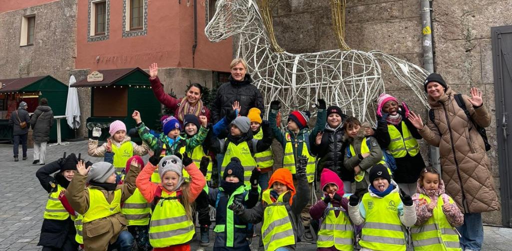 Stephanie Knabl Eine Gruppe von Kindern in leuchtend gelben Sicherheitswesten posiert gemeinsam mit drei Erwachsenen für ein fröhliches Gruppenfoto auf einem Platz vor historischen Gebäuden. Hinter ihnen steht eine große, kunstvolle Drahtskulptur eines Reiters auf einem Pferd, weihnachtlich dekoriert.