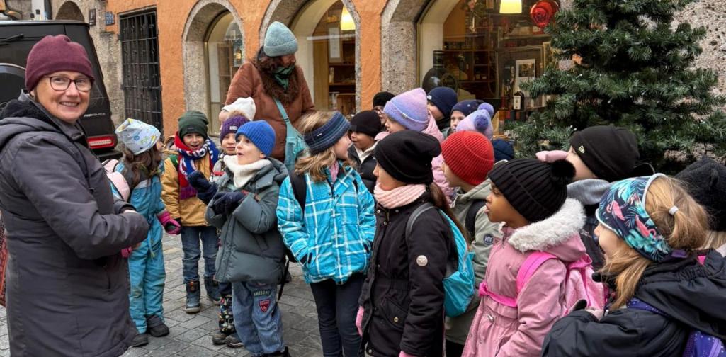 Alexandra Sigitz Eine Gruppe von Kindern in Winterkleidung steht in einer engen Altstadtgasse vor einem orangefarbenen Gebäude mit der Aufschrift „Rauch Mehlniederlage“. Eine Erwachsene spricht zu den Kindern, die aufmerksam zuhören. Neben ihnen steht ein kleiner geschmückter Weihnachtsbaum, und im Hintergrund leuchten die Schaufenster eines Geschäfts.