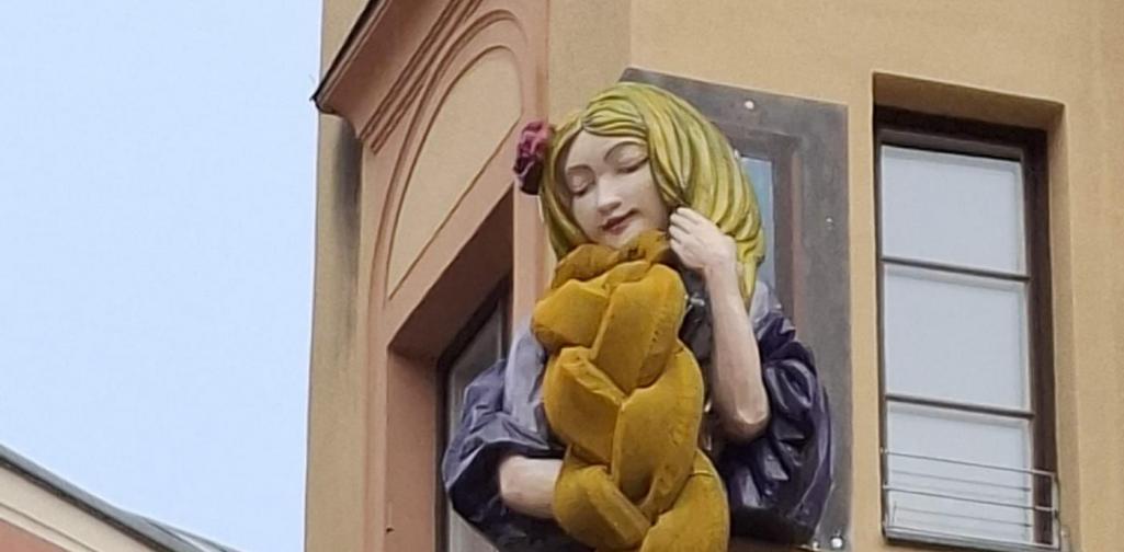 Alexandra Sigitz Eine große Rapunzel-Figur ist an einer Gebäudeecke angebracht. Sie lehnt aus einem Fenster und hält einen langen, dicken, goldgelben Zopf, der mehrere Meter an der Fassade hinunterhängt. Die Figur hat lange blonde Haare, geschlossene Augen und trägt ein violettes Kleid.