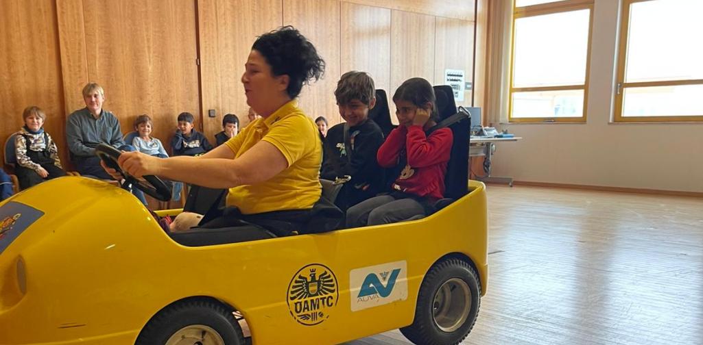 Kinder sitzen in einem gelben Übungsauto bei einer Verkehrssicherheitsaktion in einem Innenraum, betreut von Erwachsenen.
