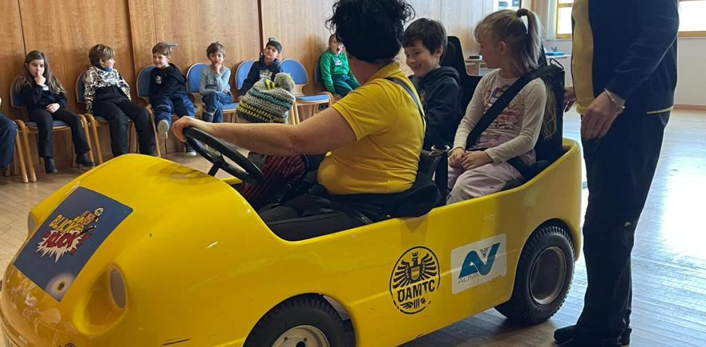 Kinder sitzen in einem gelben Übungsauto bei einer Verkehrssicherheitsaktion in einem Innenraum, betreut von Erwachsenen.