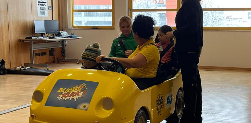 Kinder sitzen in einem gelben Übungsauto bei einer Verkehrssicherheitsaktion in einem Innenraum, betreut von Erwachsenen.