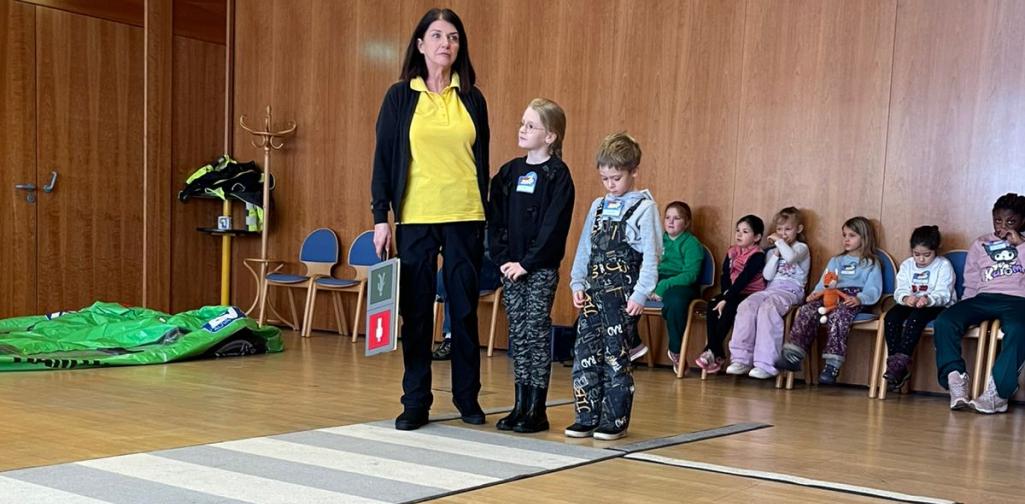 Eine Erwachsene steht mit zwei Kindern auf einem Übungs-Zebrastreifen in einem großen Raum. Weitere Kinder sitzen im Hintergrund und beobachten eine Verkehrssicherheitsübung.
