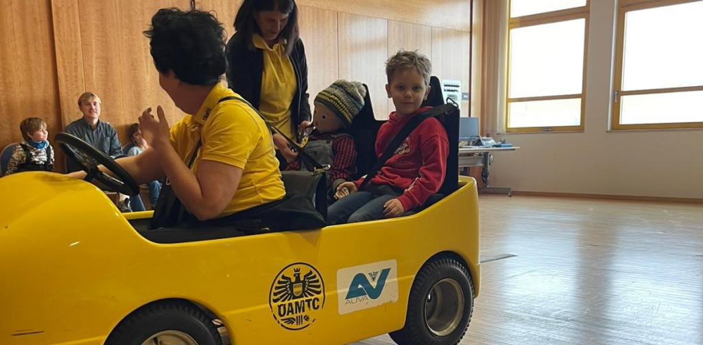 Kind sitzt in einem gelben Übungsauto bei einer Verkehrssicherheitsaktion in einem Innenraum, betreut von Erwachsenen.