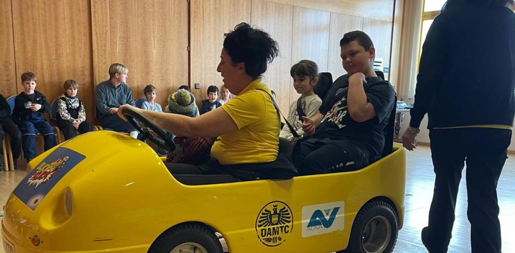 Kinder sitzen in einem gelben Übungsauto bei einer Verkehrssicherheitsaktion in einem Innenraum, betreut von Erwachsenen.