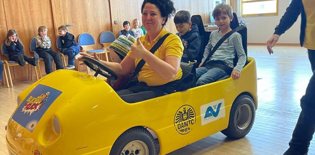 Kinder sitzen in einem gelben Übungsauto bei einer Verkehrssicherheitsaktion in einem Innenraum, betreut von Erwachsenen.