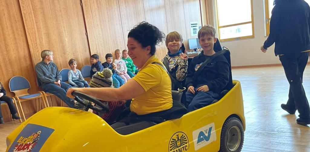 Kinder sitzen in einem gelben Übungsauto bei einer Verkehrssicherheitsaktion in einem Innenraum, betreut von Erwachsenen.