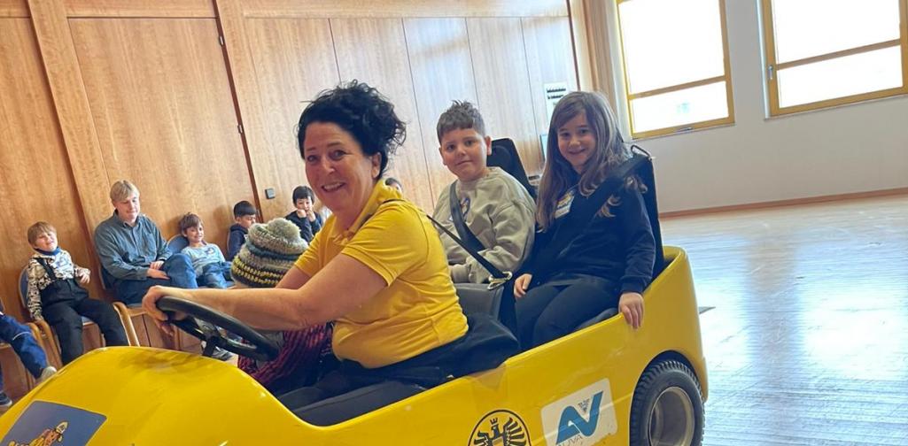 Kinder sitzen in einem gelben Übungsauto bei einer Verkehrssicherheitsaktion in einem Innenraum, betreut von Erwachsenen.