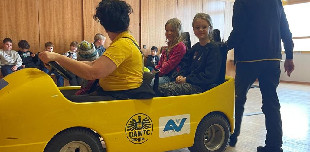 Kinder sitzen in einem gelben Übungsauto bei einer Verkehrssicherheitsaktion in einem Innenraum, betreut von Erwachsenen.