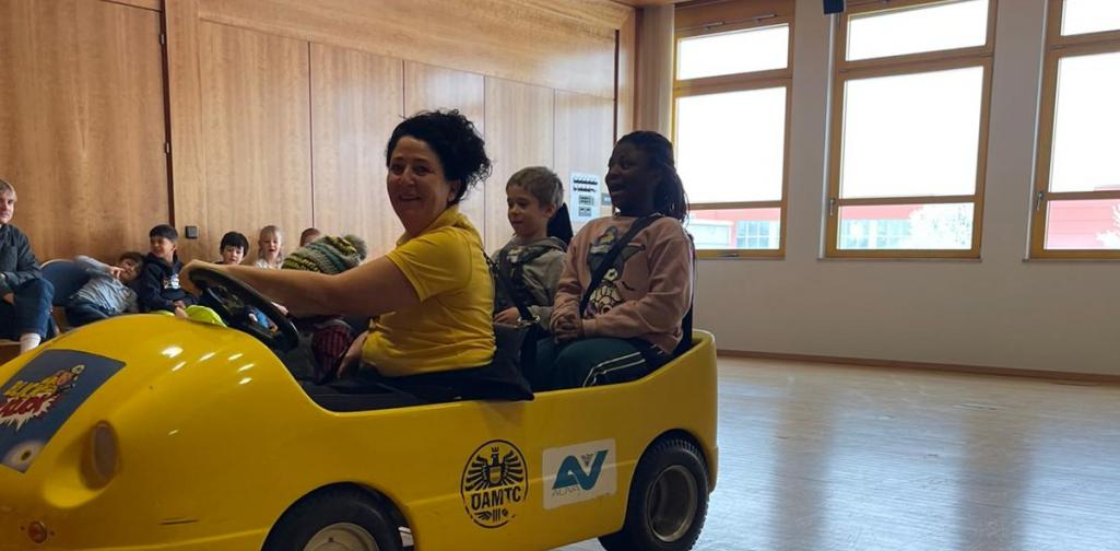 Kinder sitzen in einem gelben Übungsauto bei einer Verkehrssicherheitsaktion in einem Innenraum, betreut von Erwachsenen.