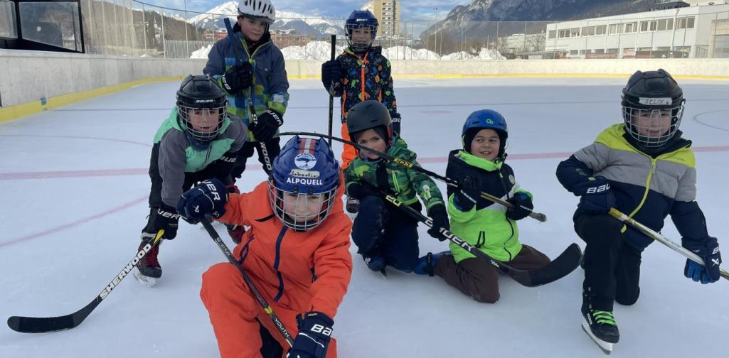Kinder einer Schulgruppe beim Eislaufen im Freien: Sie knien mit Helmen und Eishockeyschlägern auf der Eisfläche, Berge im Hintergrund.