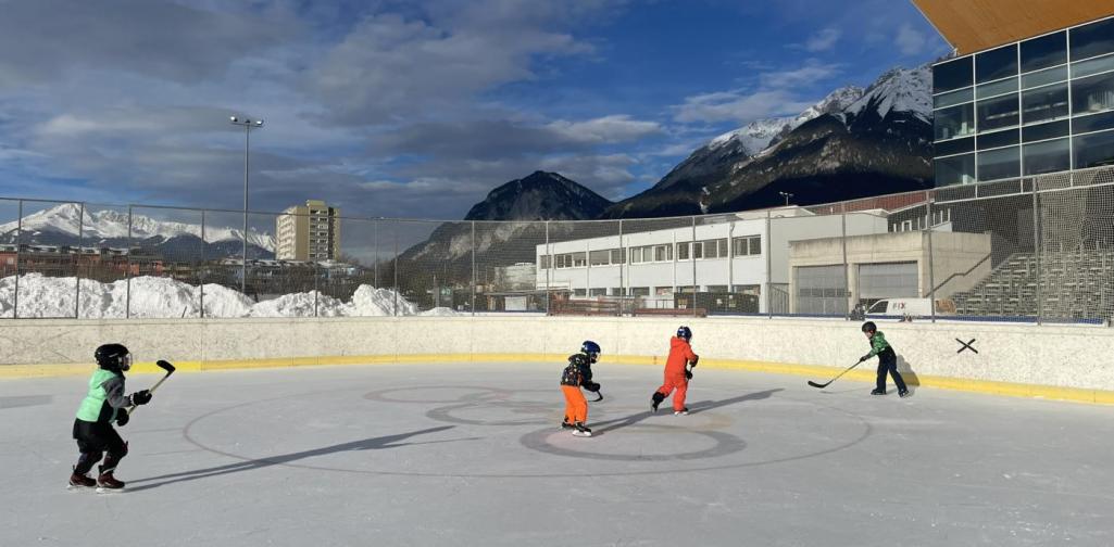 Vier Kinder in Winterkleidung und mit Helmen spielen mit Eishockeyschlägern auf einer Eisfläche im Freien. Die Eisbahn ist von einer Bande umgeben, dahinter sind verschneite Berge, Gebäude und ein blauer Himmel sichtbar.