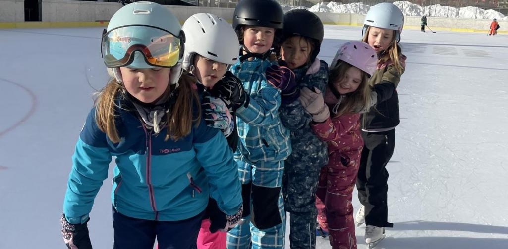 Sechs Kinder in Winterkleidung und mit Helmen stehen hintereinander auf Schlittschuhen auf einer Eisfläche; im Hintergrund sind Berge zu sehen.
