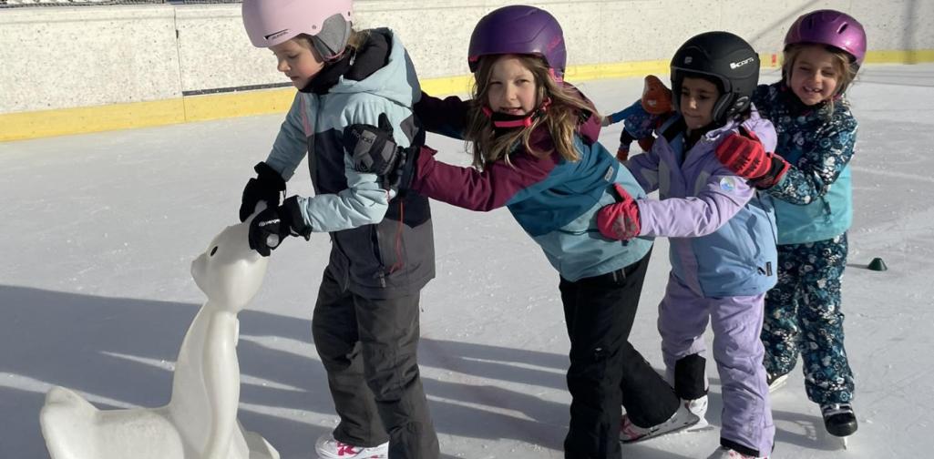 Fünf Kinder in Winterkleidung und mit Helmen fahren auf Schlittschuhen hintereinander über eine Eisfläche. Das vordere Kind schiebt eine Eislaufhilfe, die anderen halten sich gegenseitig fest.