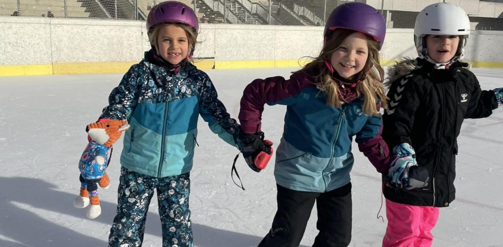 Kinder einer Schulgruppe üben gemeinsam das Schlittschuhlaufen, indem sie sich auf der Eisfläche an den Händen halten.